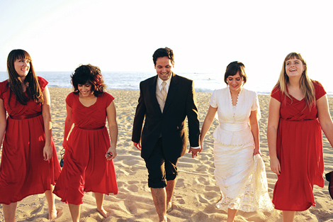 The bridal party, wearing bright red dresses, walk with the bride and groom on the beach following their wedding in Los Angeles
