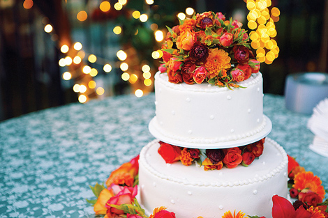 This white wedding cake is adorned with colorful flowers, including red roses, and bright orange and maroon flowers