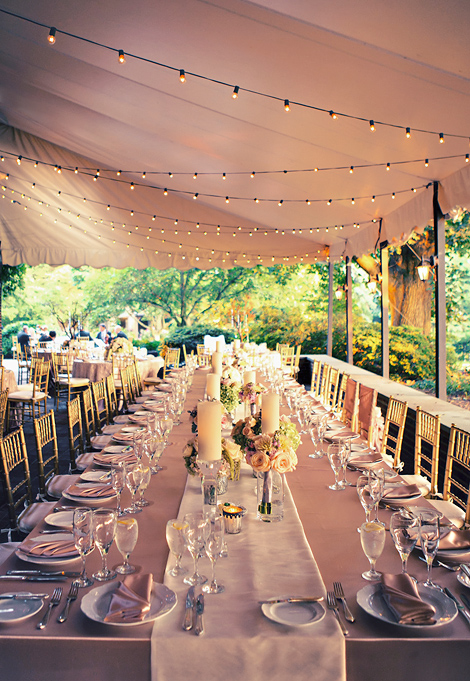 The bridal party table is set with soft pink linens and pink roses, candles and the tent is elegantly strung with lights to create a romantic mood