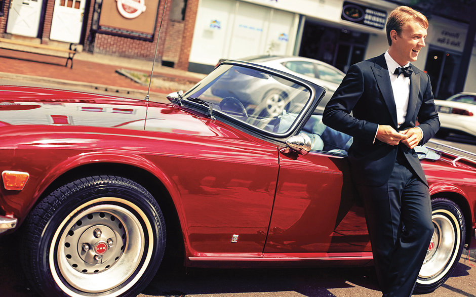 The groom leans against a vintage red convertible car before the wedding