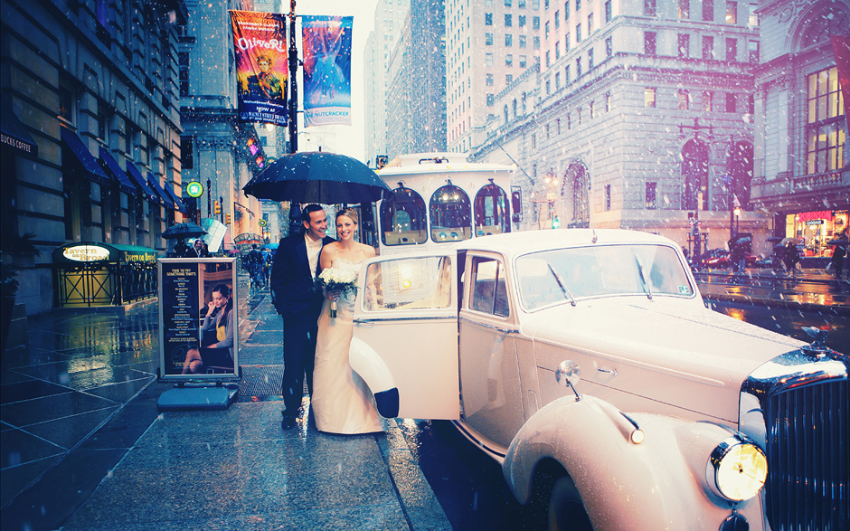 The bride and groom pose on Broad Street in Philadelphia as the snow falls, for their winter wedding, posing next to the vintage antique car, photography by Peter Van Beever