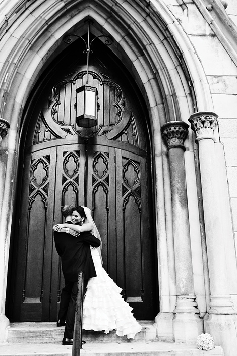 The bride and groom hug on the steps outside the church doors before their Philadelphia wedding, the bride is wearing a veil and her bouquet is on the steps.