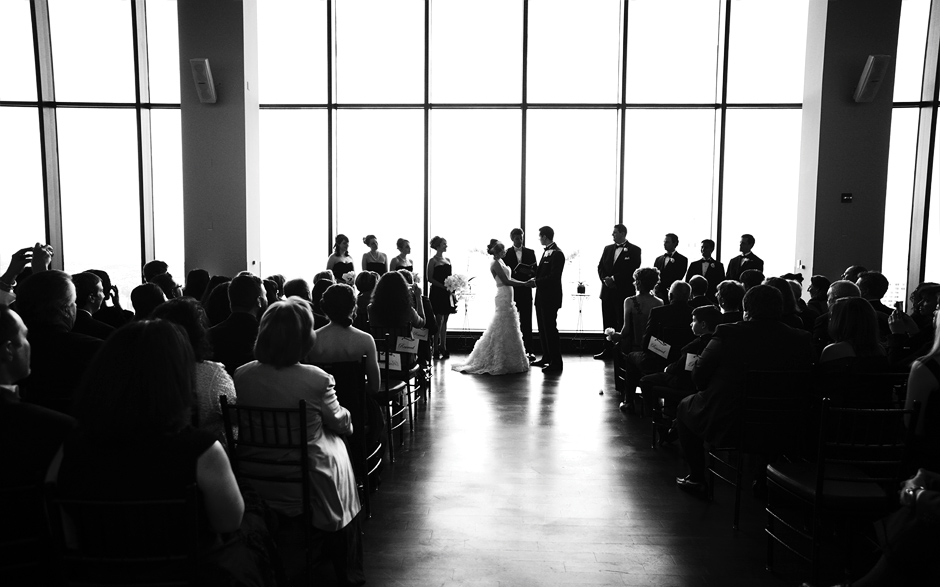The bride and groom exchange vows in front of friends and family, looking beautiful and elegant in front of the floor to ceiling windows at the State Room in Boston, photography by Peter Van Beever