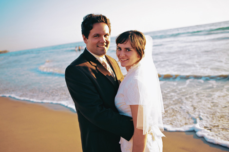 The bride and groom pose for a photo on the beach, with the blue ocean foaming at their feet after their Los Angels wedding