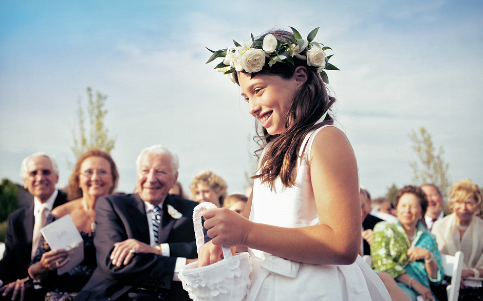 The flower girl walks down the aisle carrying a silky white basket with a ring of flowers in her hair, smiling, at the Belle Mer Newport Rhode Island